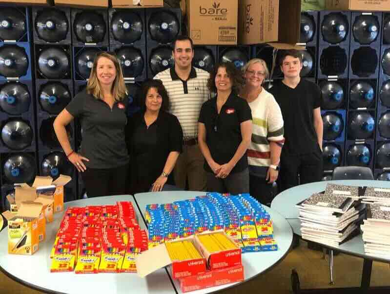Six adults and one teen stand behind tables filled with school supplies, including pencils, crayons, glue sticks, and notebooks, ready for donation.