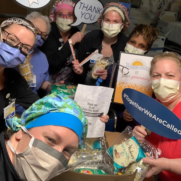 Masked healthcare workers smiling and holding thank-you signs around a donation box filled with snacks and Cow Tales candy.