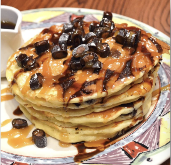 A stack of Caramel Brownie Cow Tales pancakes on a plate, topped with caramel and chocolate sauce, sprinkled with pieces of Caramel Brownie Cow Tales candy, and served with syrup on the side.