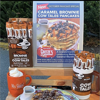 Promotional display for Caramel Brownie Cow Tales Pancakes, featuring a sign with a stack of pancakes topped with syrup and chocolate pieces. The display includes containers filled with Cow Tales candies, a plate of pancakes with chocolate and whipped cream, and a Miss Shirley's Cafe mug with whipped cream. The sign notes it's an October special in partnership with Goetze's Candy Company.