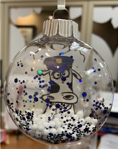 A clear Christmas ornament filled with white beads and blue glitter, featuring Duke the cow wearing a hat.