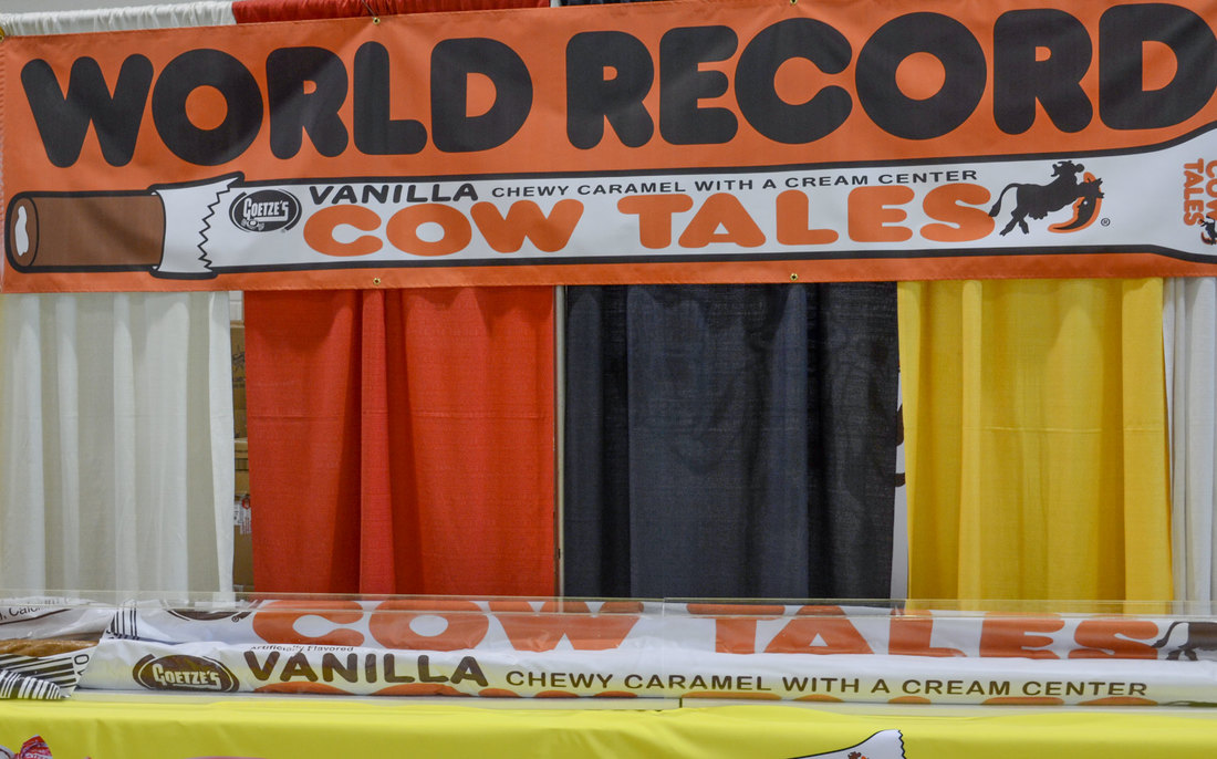 A large banner reading “World Record” and advertising Goetze’s Vanilla Cow Tales chewy caramel with a cream center is displayed at an indoor event. Below the banner, a giant replica of a Cow Tales candy stick is encased in a clear display box. The backdrop consists of red, black, yellow, and white curtains.