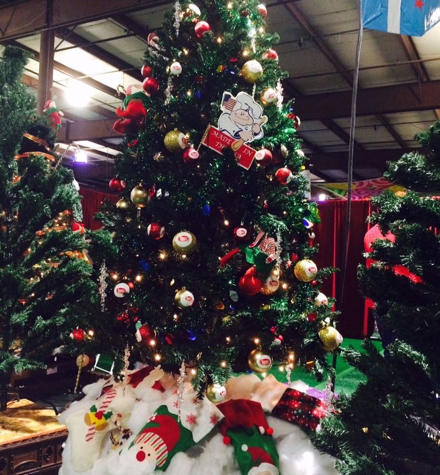 A decorated Christmas tree at an indoor event, featuring lights, ornaments with the Goetze’s logo, gold and red baubles, and a “Made in the USA” sign with a cartoon cow. Stockings and cotton snow surround the base.