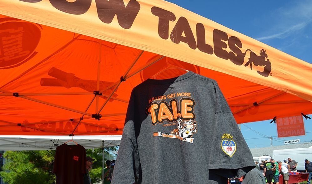 A dark gray T-shirt displayed under an orange Cow Tales-branded canopy tent at an outdoor event. The T-shirt reads “Pullers Get More Tale” with a cartoon cow holding a Cow Tales candy stick. The sleeve has a patch that says “A Proud Sponsor of NTPA.” In the background, people are gathered near booths and white tents under a clear blue sky.