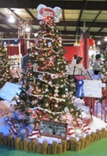 A decorated Christmas tree at an indoor event, featuring lights, ornaments with the Goetze’s logo, gold and red baubles, and a “Made in the USA” sign with a cartoon cow. Stockings and cotton snow surround the base.